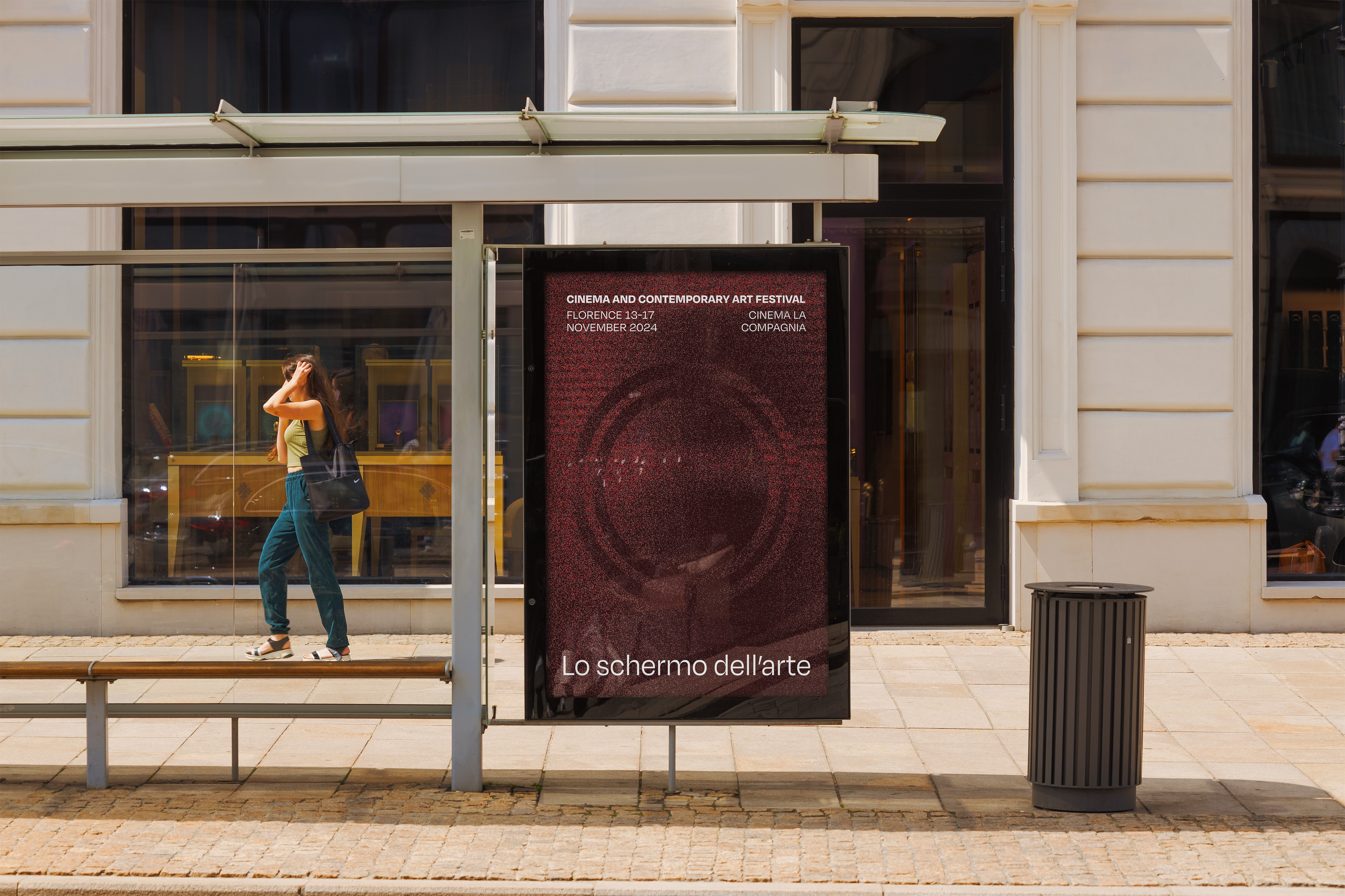 A bus stop with a red poster displayed at it. A woman walks through the background.